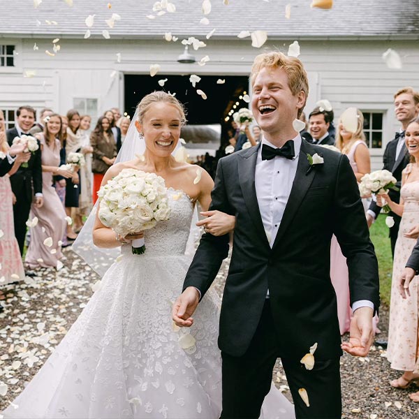 A joyful bride and groom walk arm in arm outside, smiling as guests throw flower petals. The bride holds a white bouquet and wears a lace gown; the groom wears a black tuxedo. Friends and family celebrate around them.