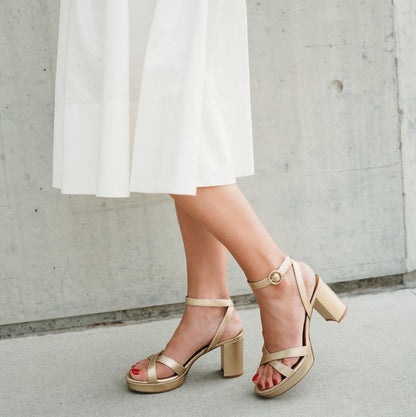 Beige sandals worn with a white skirt against a concrete wall.