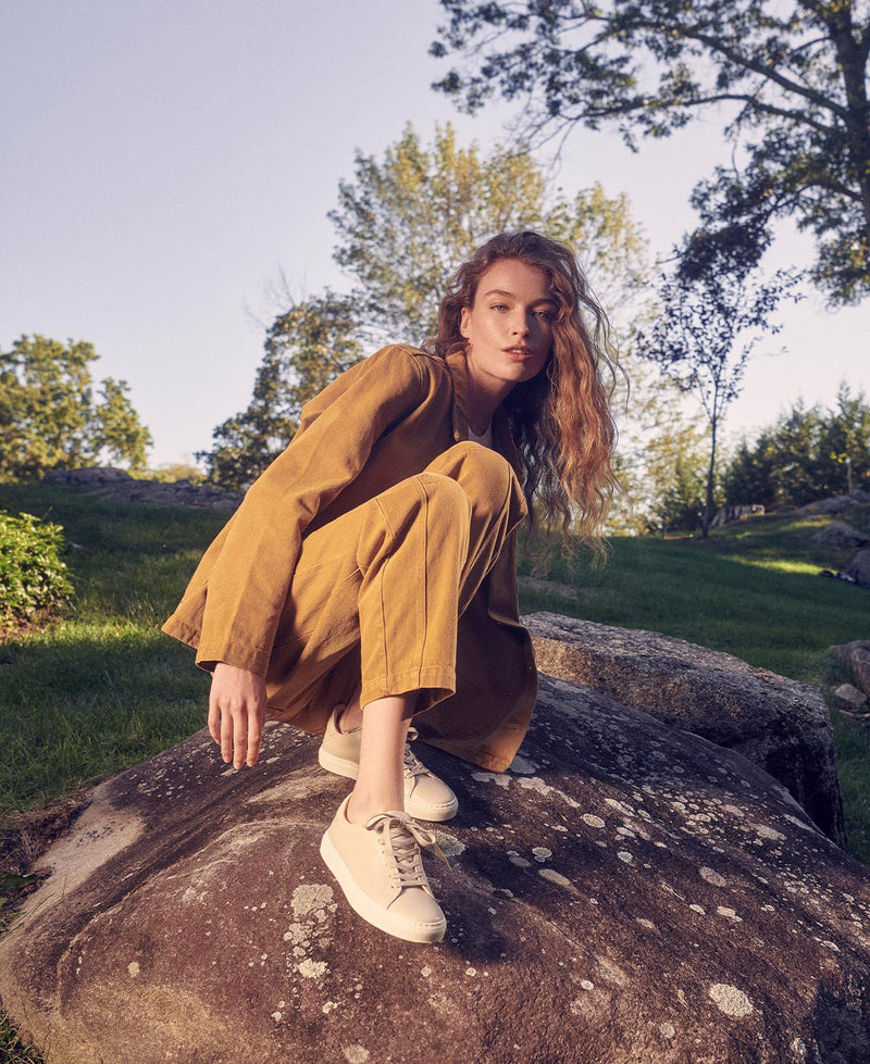 A woman with long wavy hair, dressed in a mustard yellow suit and cream sneakers, crouches on a large rock outdoors surrounded by grass, trees, and sunlight.