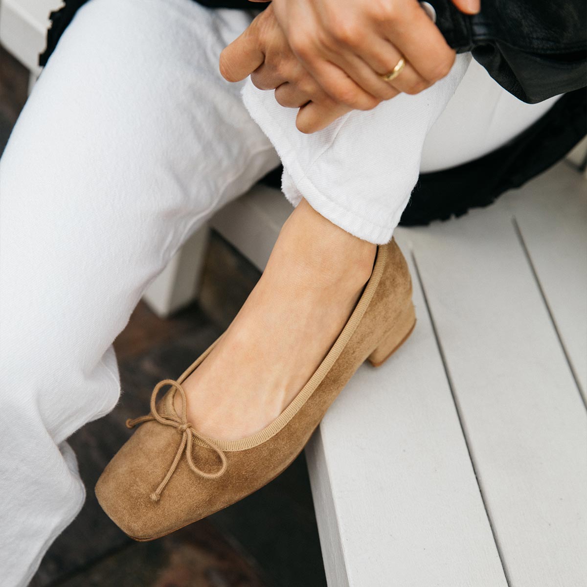 A person in white pants and a black jacket sits on a white bench, adjusting their hem to showcase The Ada - Biscotti Suede by margaux-development: a tan suede low-heeled ballet pump with sacchetto construction and a bow.