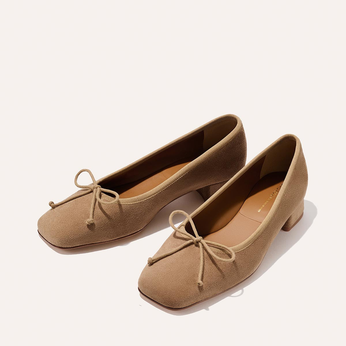 The Ada - Biscotti Suede by margaux-development features classic light brown suede ballet pumps with low heels, square toes, and front bows, showcased on a light background.