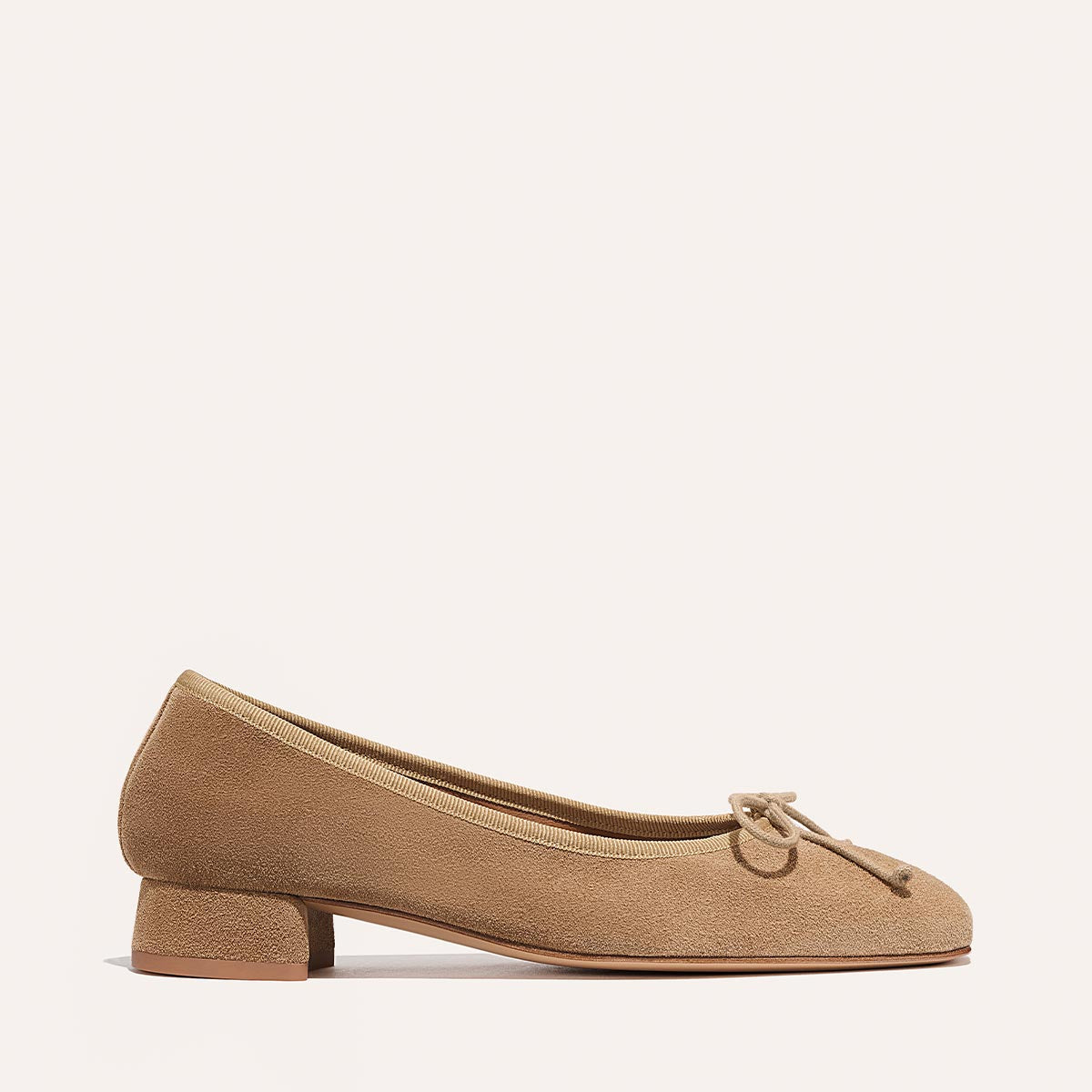 The Ada - Biscotti Suede by margaux-development is a classic tan suede ballet pump with a rounded toe, small block heel, and decorative front bow, shown against an off-white background.