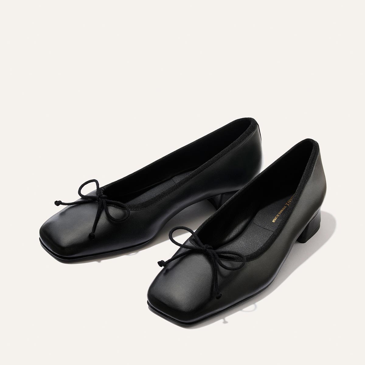 A pair of The Ada - Black Nappa ballet pumps by Margaux, featuring low heels, square toes, and elegant bow details, crafted with refined sacchetto construction and displayed on a light background.
