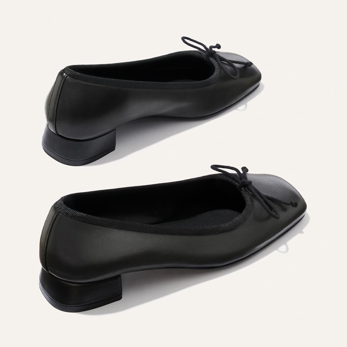 Margaux’s The Ada - Black Nappa flats, crafted in a classic ballet pump silhouette with low block heels and small front bows, are shown from the side against a white background.