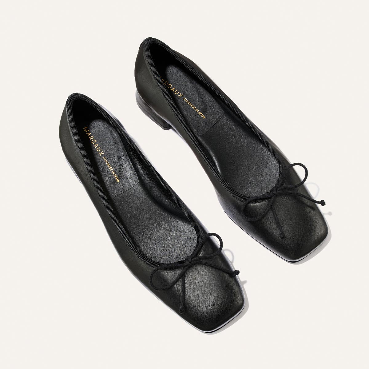 A pair of Margaux The Ada - Black Nappa ballet flats with square toes and small bows on the front, crafted with sacchetto construction for enhanced comfort, displayed on a light background.