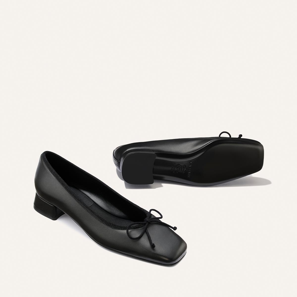 A pair of The Ada flats by Margaux in black nappa leather, featuring low square block heels and bow accents. One shoe is positioned upright to showcase the side and front, while the other lies on its side, highlighting the sole and heel. Designed with sacchetto construction for enhanced flexibility and comfort.