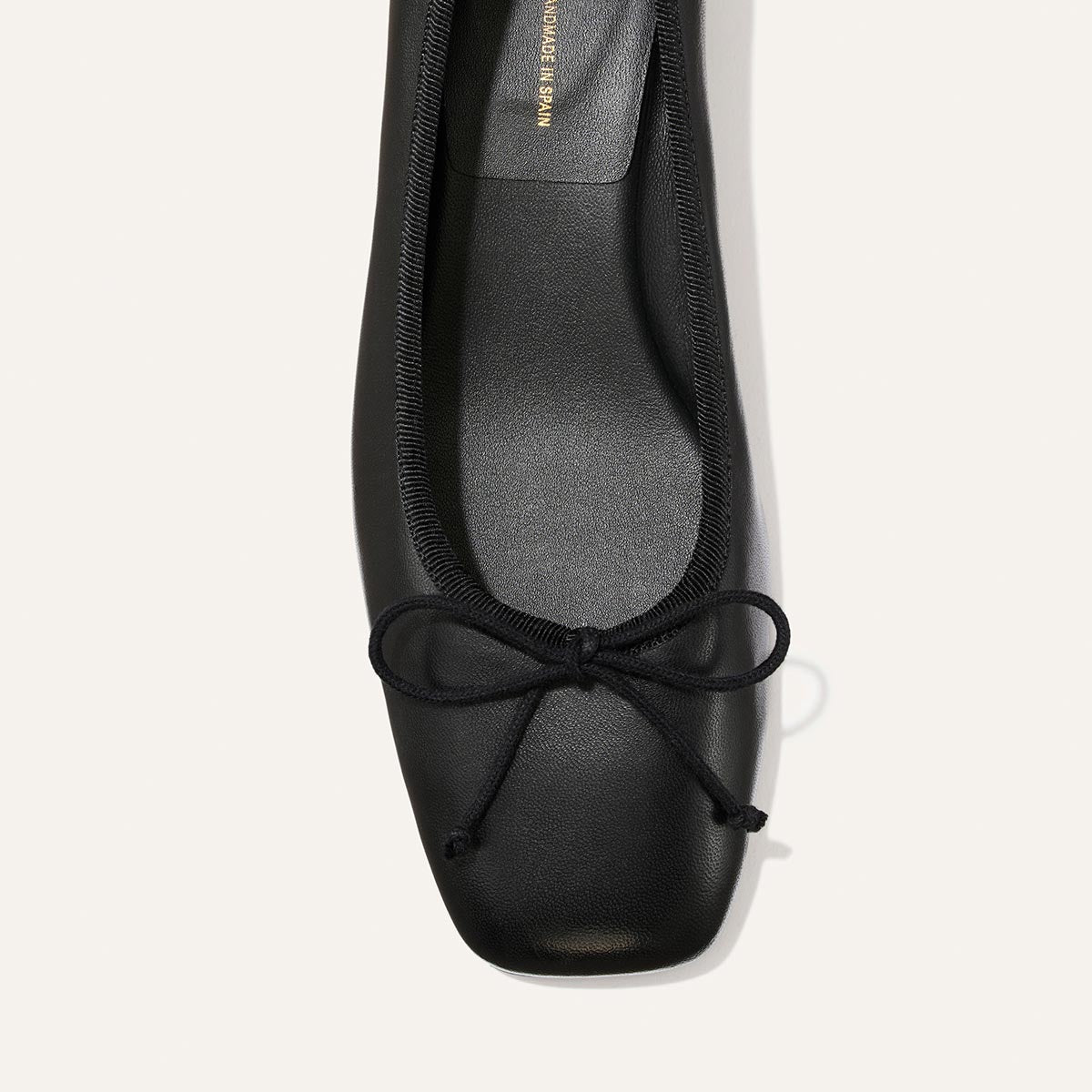 Top view of The Ada - Black Nappa by Margaux, a black leather ballet pump featuring a square toe and a petite black bow on the front. Crafted with sacchetto construction, its interior is also black, and it is photographed against a plain light background.