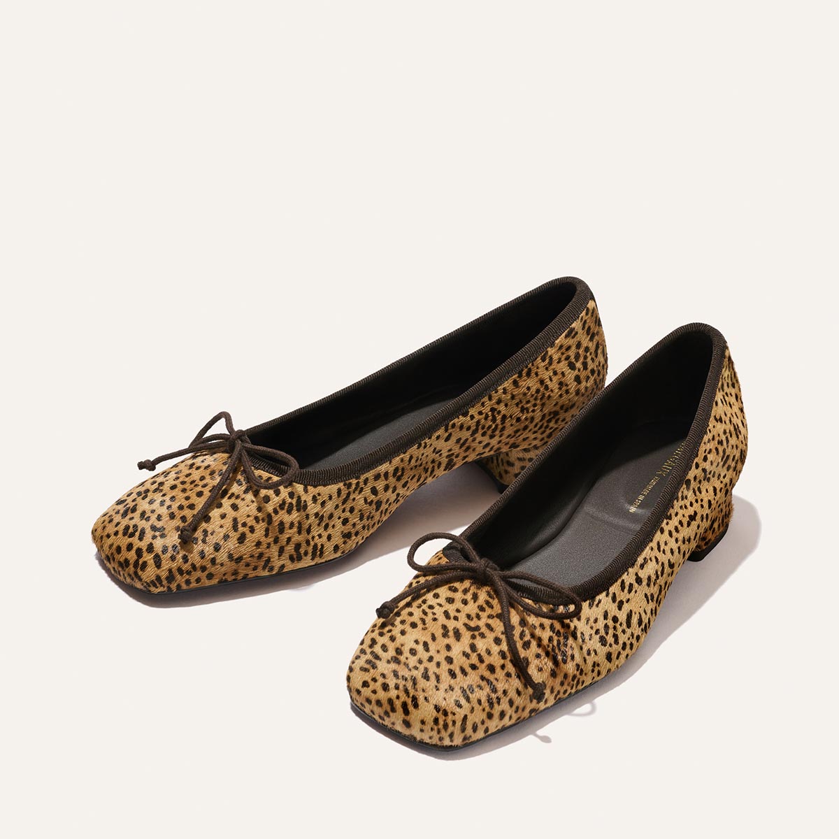 A pair of The Ada - Tan Mini Cheetah Haircalf ballet flats by Margaux, featuring a cheetah-print design with small black bows on the front, displayed on a white background.