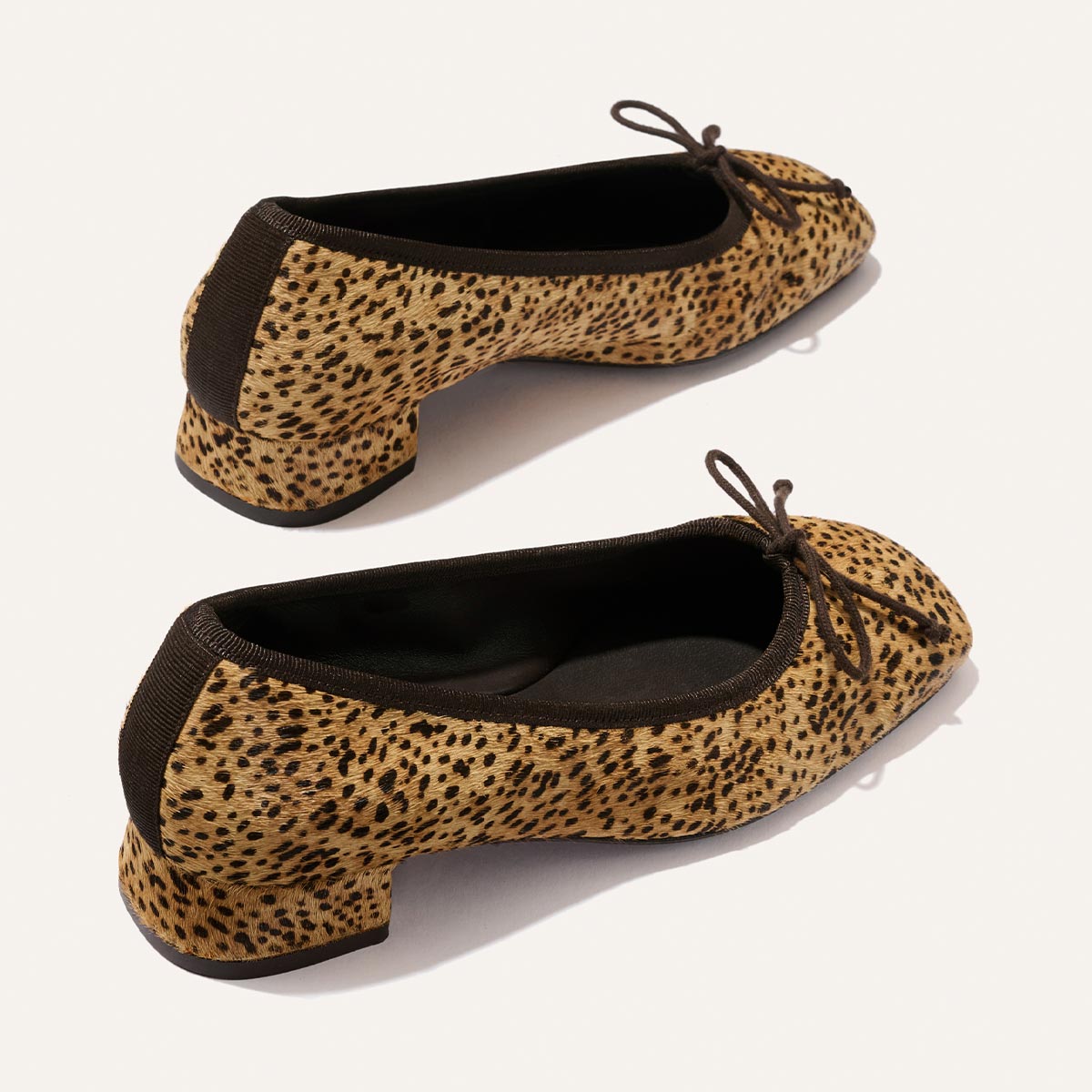 Two “The Ada - Tan Mini Cheetah Haircalf” ballet flats by Margaux, featuring brown trim and bows, are displayed on a light background, showcasing both the side and back views of the shoes.
