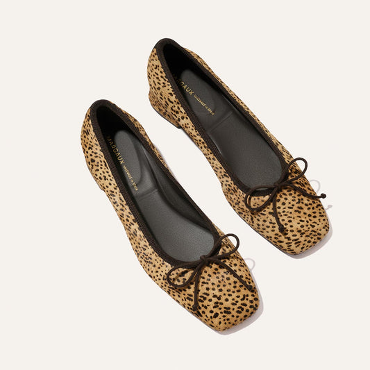 A pair of Margaux’s “The Ada” flats in tan mini cheetah haircalf, featuring black trim and black bow accents, displayed on a plain white background.