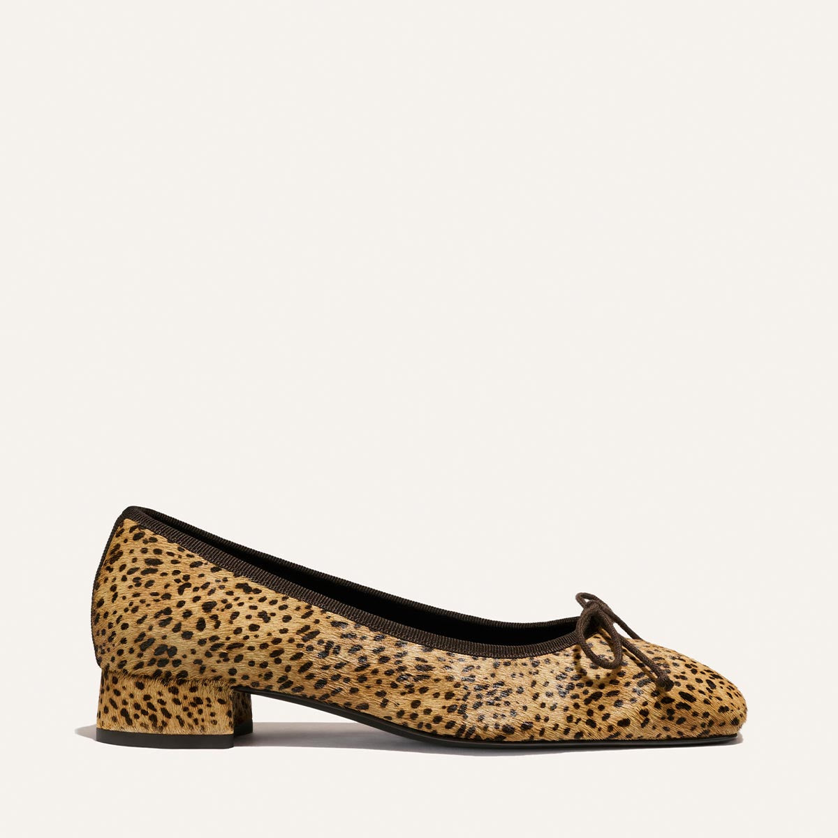 The Ada - Tan Mini Cheetah Haircalf by Margaux is a ballet flat in a cheetah-print haircalf with black trim, a low block heel, and a small black bow on the toe, photographed against a plain light background.
