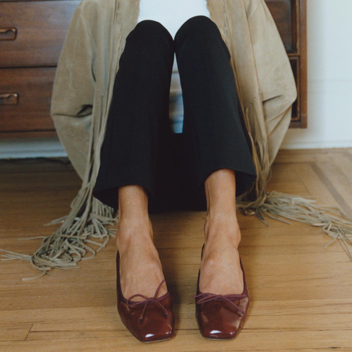 A person sits on a wooden floor in front of a dresser, wearing black pants, a fringed beige jacket, and The Ada - Claret Crinkle Patent ballet pumps by margaux-development. Only the lower body is visible.