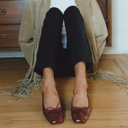 A person sits on a wooden floor in front of a dresser, wearing black pants, a fringed beige jacket, and The Ada - Claret Crinkle Patent ballet pumps by margaux-development. Only the lower body is visible.