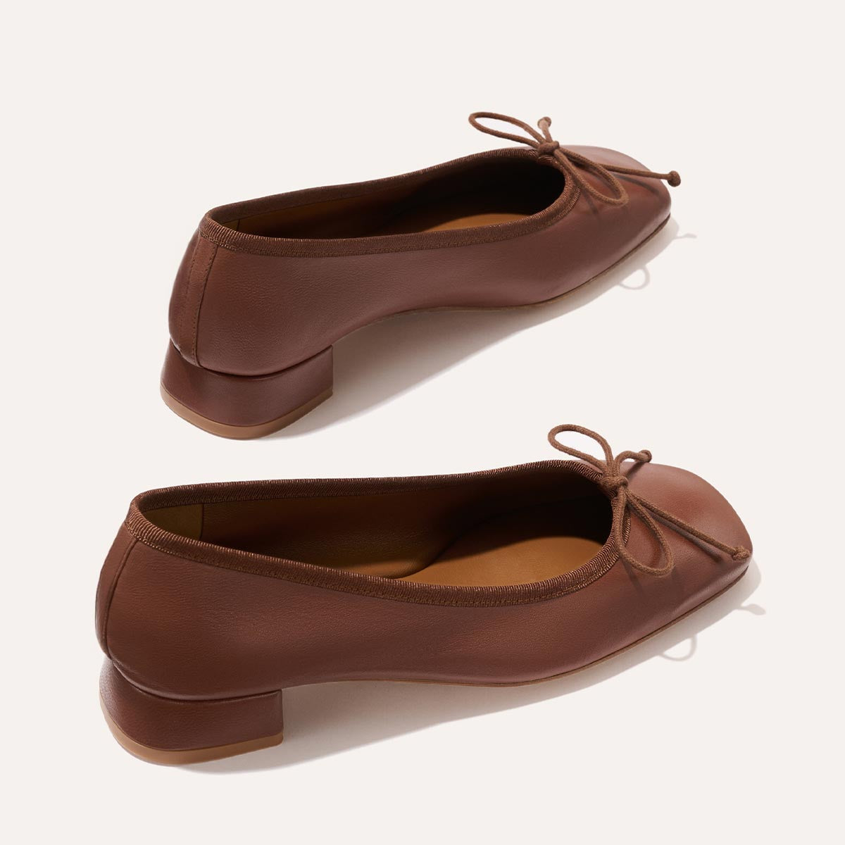 The Ada - Cognac Nappa by margaux-development: brown leather ballet pumps with low block heels, rounded toes, sacchetto construction for a glove-like fit, and thin bow ties on the front, shown on a light background.