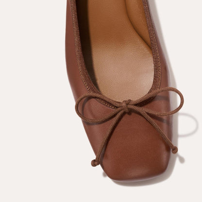 Close-up of The Ada - Cognac Nappa ballet pump by margaux-development: brown leather with square toe, trimmed edges, matching bow, tan insole, and sacchetto construction for a glove-like fit against an off-white background.