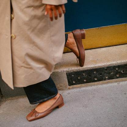 A person in a beige trench coat and dark jeans stands on a sidewalk, wearing margaux-developments The Ada - Cognac Nappa ballet pumps. Behind them is a blue door highlighted by a gold metallic strip at the bottom.