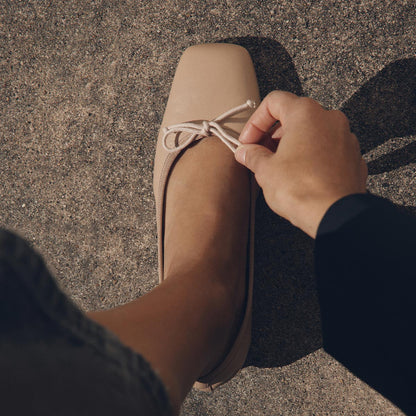 A person adjusts the bow on The Ada - Dune Nappa, a nude-colored low-heeled ballet pump by Margaux, while standing on a textured concrete surface.