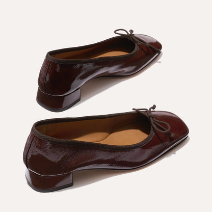 The Ada - Espresso Crinkle Patent by Margaux are glossy dark brown ballet flats with low block heels, square toes, and small front bows, crafted with sacchetto construction and shown on a light beige background.