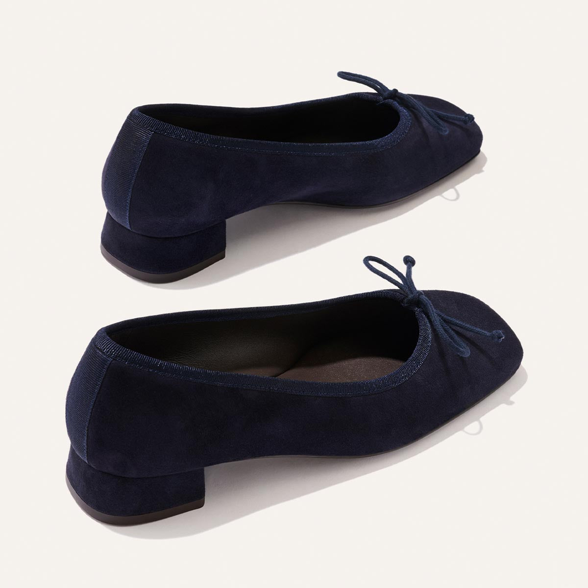 The Margaux Ada in Indigo Suede: low-heeled ballet pumps with rounded toes, small bows, and sacchetto construction for a glove-like fit, shown against a light background.