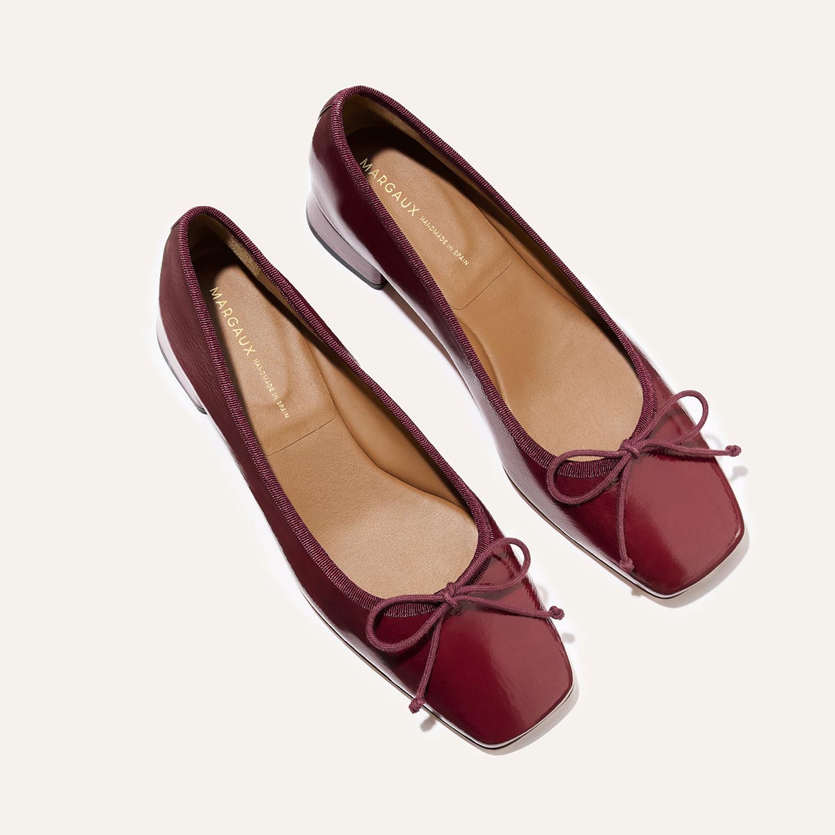 The Ada - Claret Crinkle Patent by margaux-development are low-heeled burgundy patent ballet pumps with square toes and small bows, displayed on a light background for a classic look.