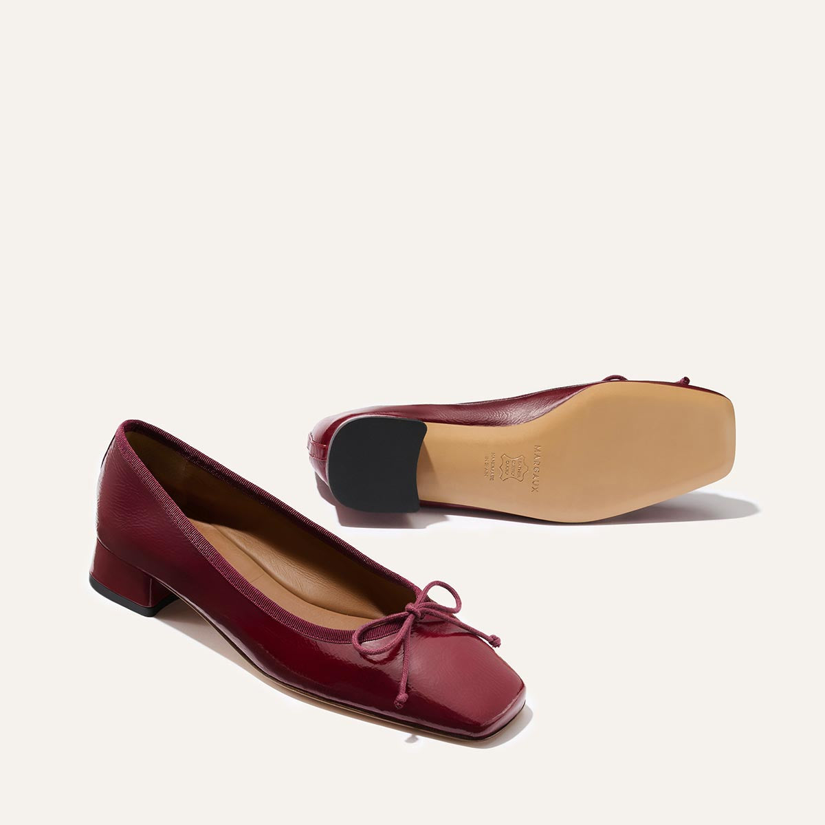 A pair of The Ada - Claret Crinkle Patent ballet flats by margaux-development, featuring square toes, bow detail, and small block heels. One shoe stands upright while the other rests on its side to reveal the sole.
