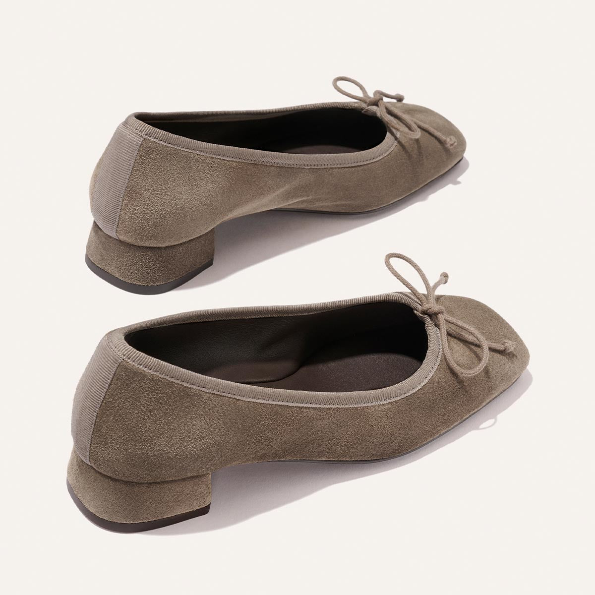 The Ada - Truffle Suede by Margaux features taupe suede ballet flats with low block heels, rounded toes, matching fabric heel trim, and small front bows. Displayed on a light background.