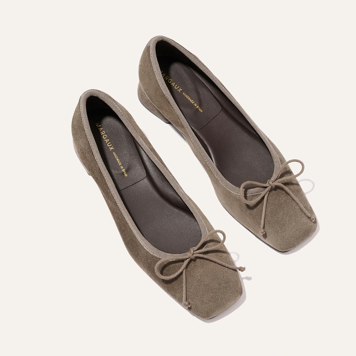 A pair of Margaux "The Ada - Truffle Suede" ballet flats featuring taupe suede, square toes, matching bows, and black insoles with gold branding are positioned diagonally on a plain off-white background.