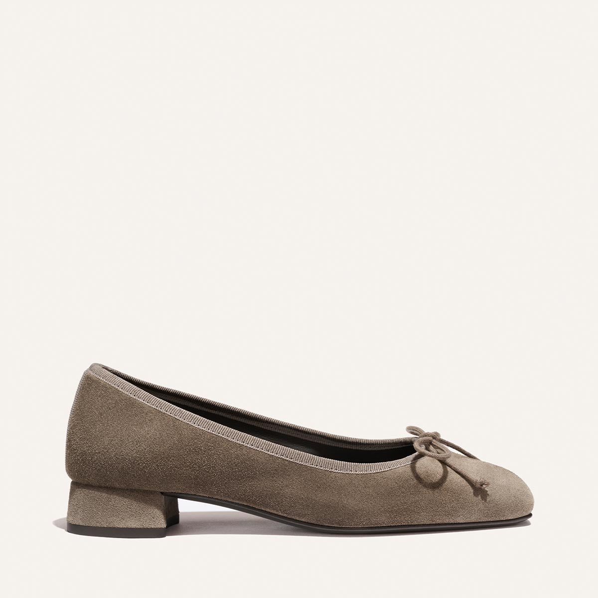 The Ada - Truffle Suede by Margaux is a taupe suede ballet flat featuring a low block heel, rounded toe, and a front bow with tassel detail. Its simple, elegant design makes it a timeless wardrobe staple.