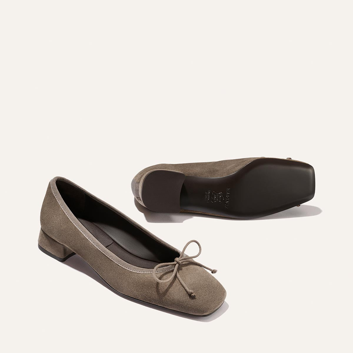 A pair of Margaux "The Ada - Truffle Suede" ballet flats in taupe suede with a small heel, bow at the toe, and black soles. One shoe displays the top and side, while the other shows the sole.