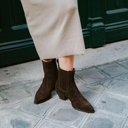 A person wearing a beige skirt and The Agnes Boot - Cocoa Suede by margaux-development stands on a stone pavement in front of a green door.
