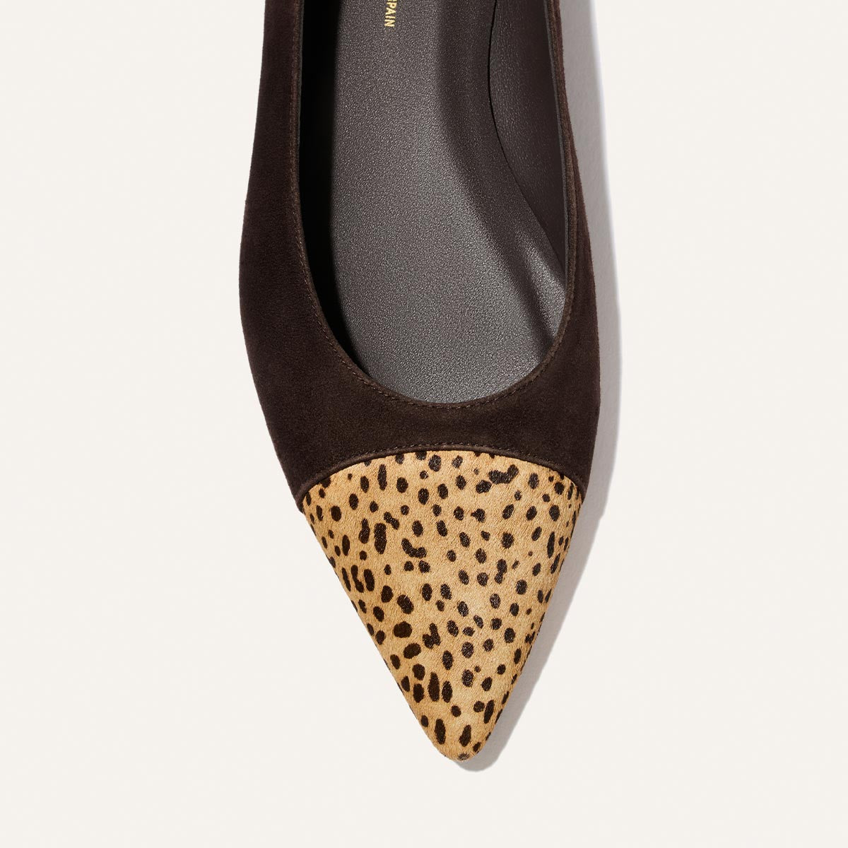 A close-up of Margaux’s The Alba flat showcases a chic pointed toe design in molasses suede, accented with a tan mini cheetah haircalf tip, set against a plain white background.