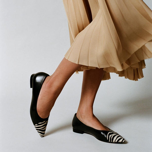A person wearing a beige pleated skirt and Margaux’s The Alba flats in Black Nappa and Zebra Haircalf stands against a plain background.