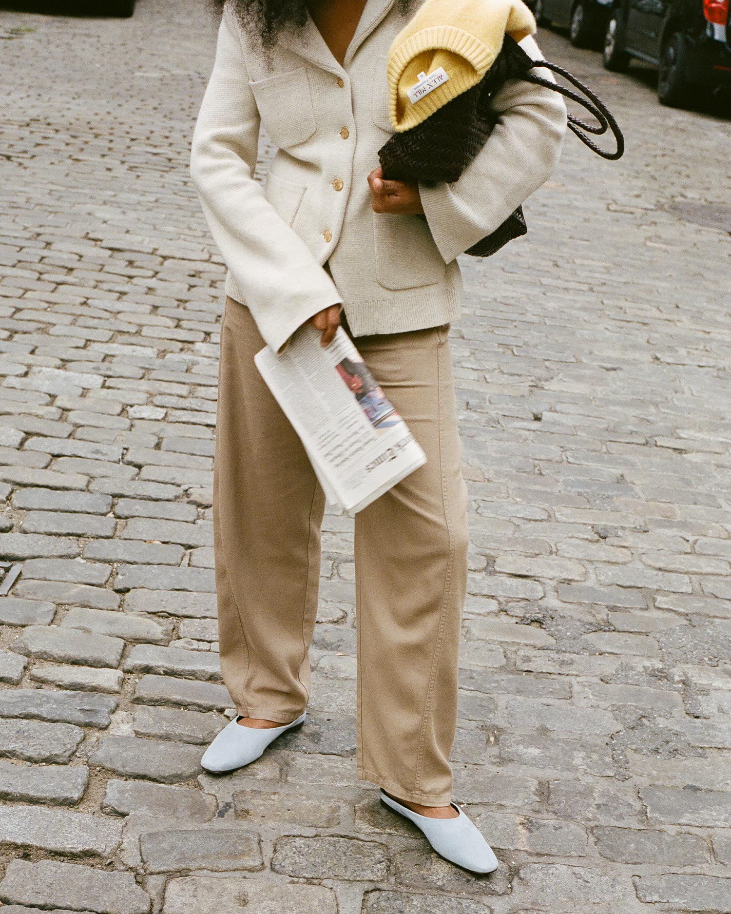 A person stands on a cobblestone street wearing a cream jacket, beige pants, and light blue shoes, holding a newspaper and a woven bag with a yellow knit hat inside. Their face is not visible.