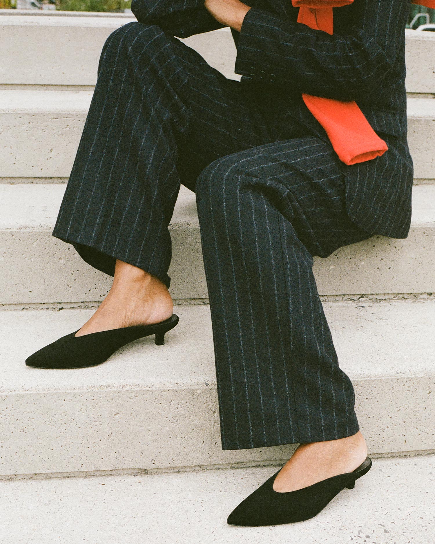 A person sits on concrete steps wearing a dark pinstripe suit, a red scarf, and black low-heeled mules. Only the lower half of their body is visible.