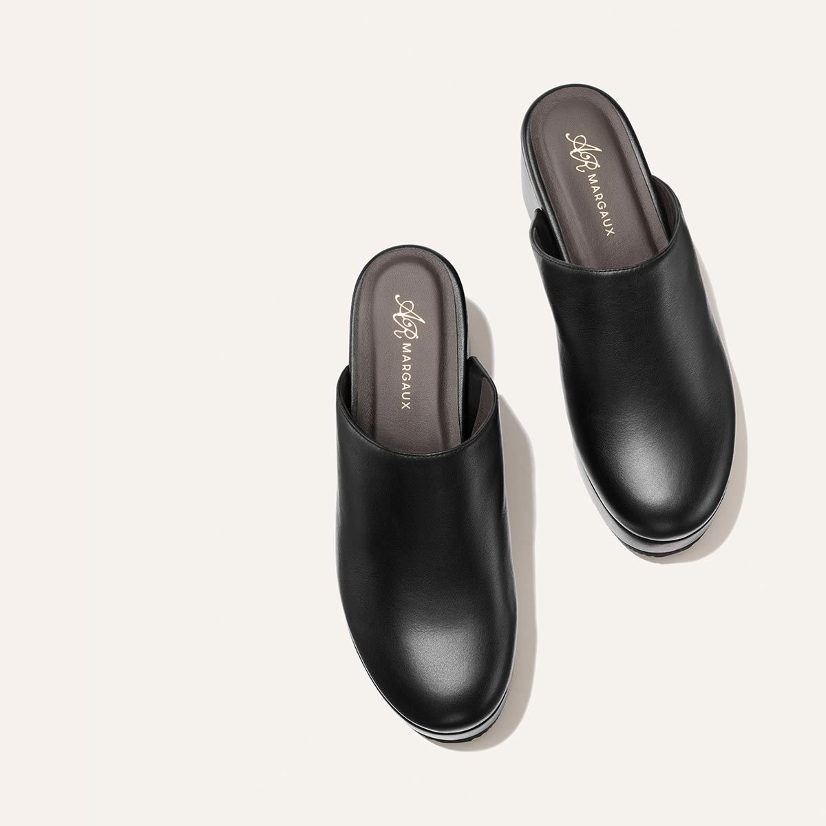 A pair of black calf leather slip-on clogs, named “The Atlantic Clog” from the Alison Roman x Margaux collection, displayed on a light background with insoles branded Margaux.