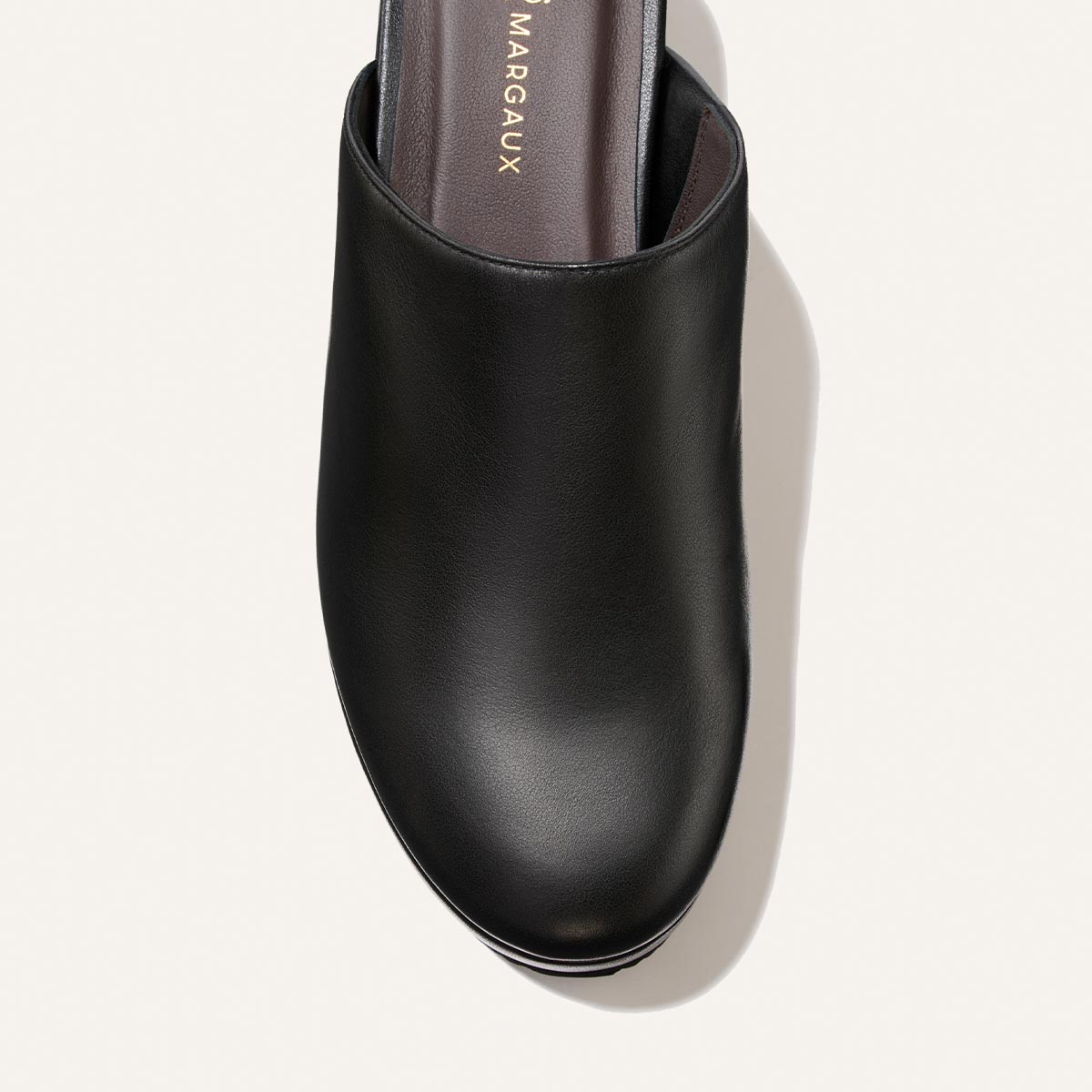 A close-up top view of the Alison Roman x Margaux: The Atlantic Clog - Black Calf slip-on mule, featuring a rounded toe and a smooth black leather upper. The Margaux brand name is partially visible on the insole, set against a clean, light background.