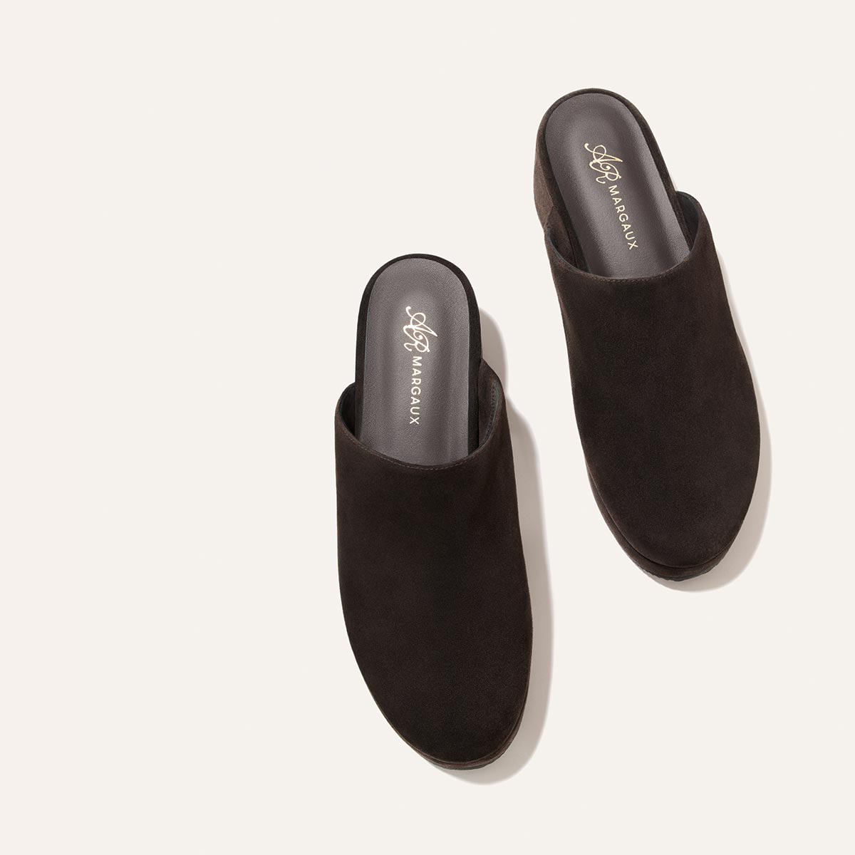 A pair of The Atlantic Clog in Dark Chocolate Suede from Margaux, designed in collaboration with Alison Roman, featuring black insoles and the Margaux logo in gold lettering, displayed on a light beige background.