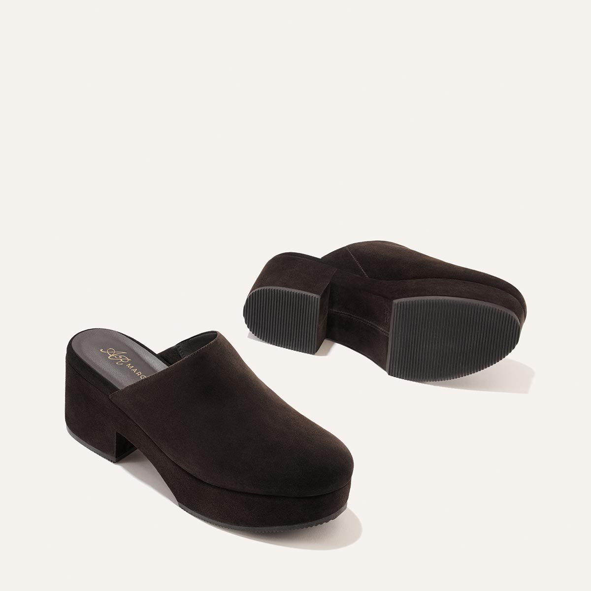 A pair of Margaux Alison Roman x Margaux: The Atlantic Clog in dark chocolate suede with chunky platform heels—one shoe stands upright while the other rests on its side to reveal the ribbed sole, all set against a simple white background.