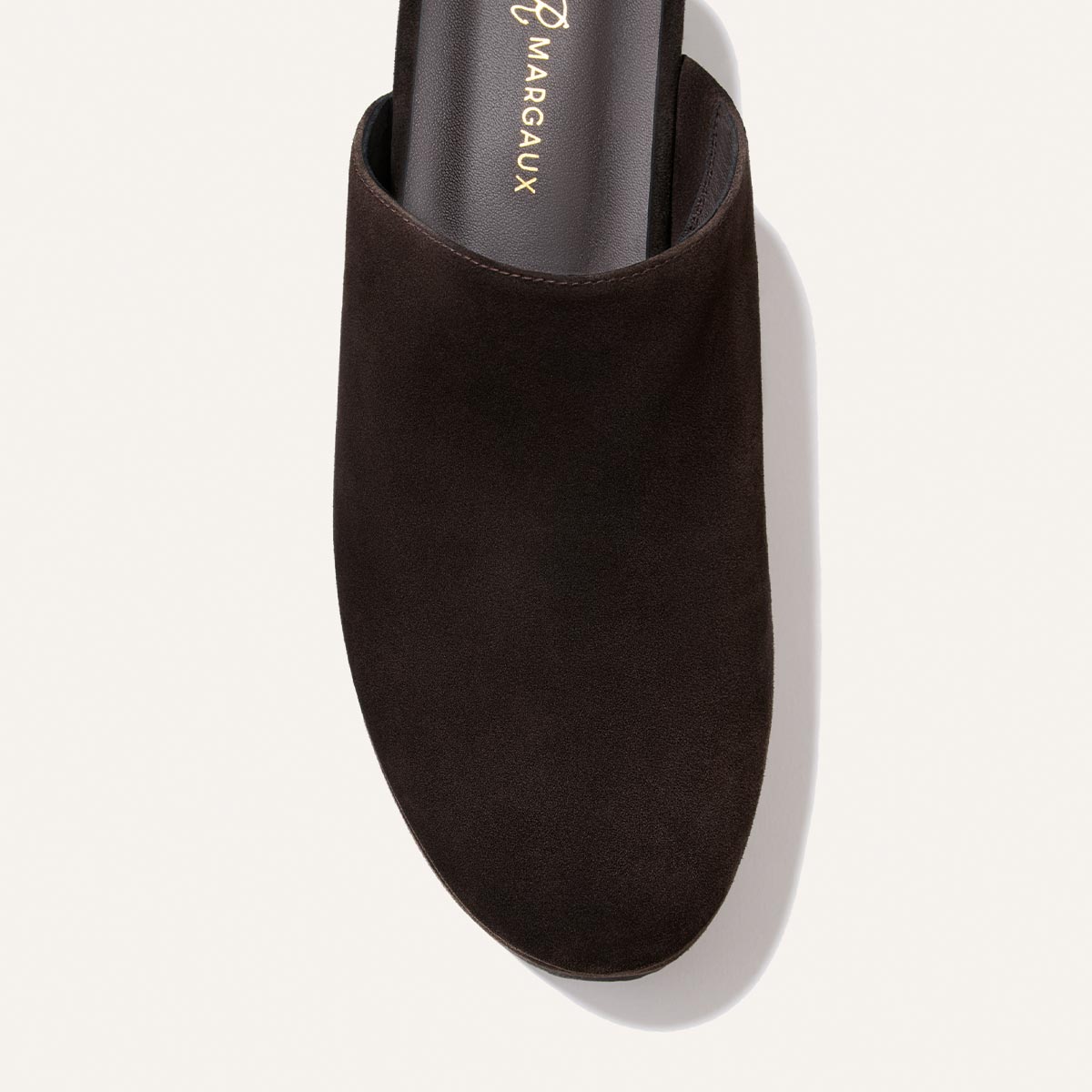 A close-up of The Atlantic Clog in dark chocolate suede from the Alison Roman x Margaux collection, featuring the Margaux brand name on the insole, photographed against a light background.