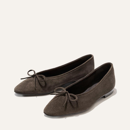 A pair of Margaux Alison Roman x Margaux: The Salamanca flats in English Brown Suede, featuring pointed toes and delicate bows on top, crafted with an unlined construction for enhanced comfort, displayed against a plain light background.