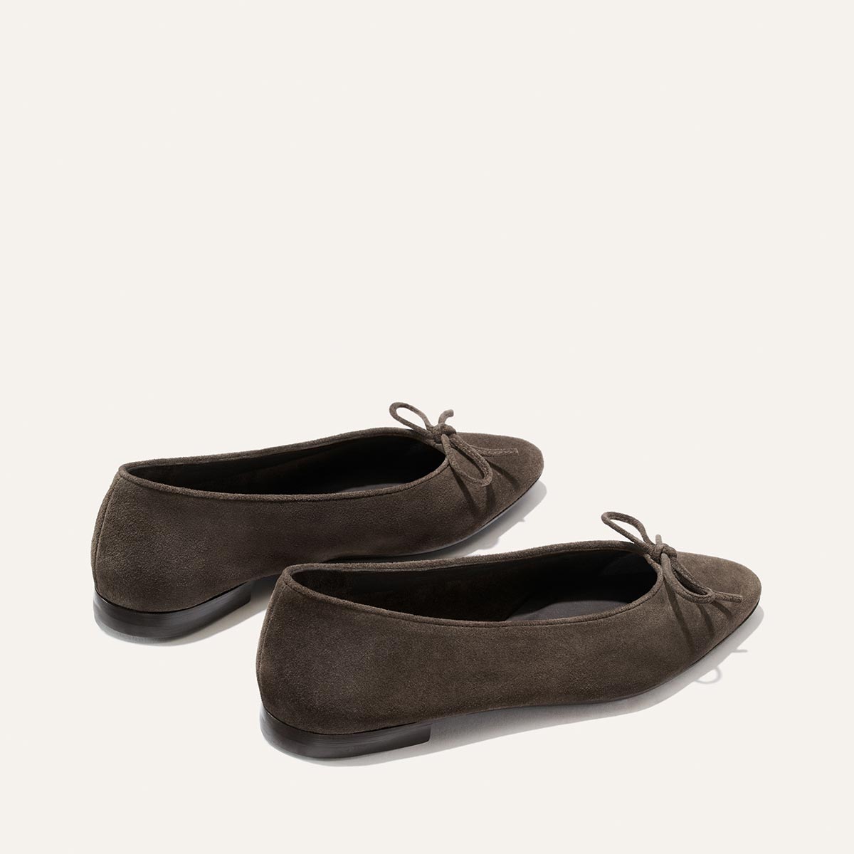 A pair of Alison Roman x Margaux: The Salamanca – English Brown Suede flats by Margaux, featuring elegant pointed toes and delicate front bows, showcased on a plain light background.