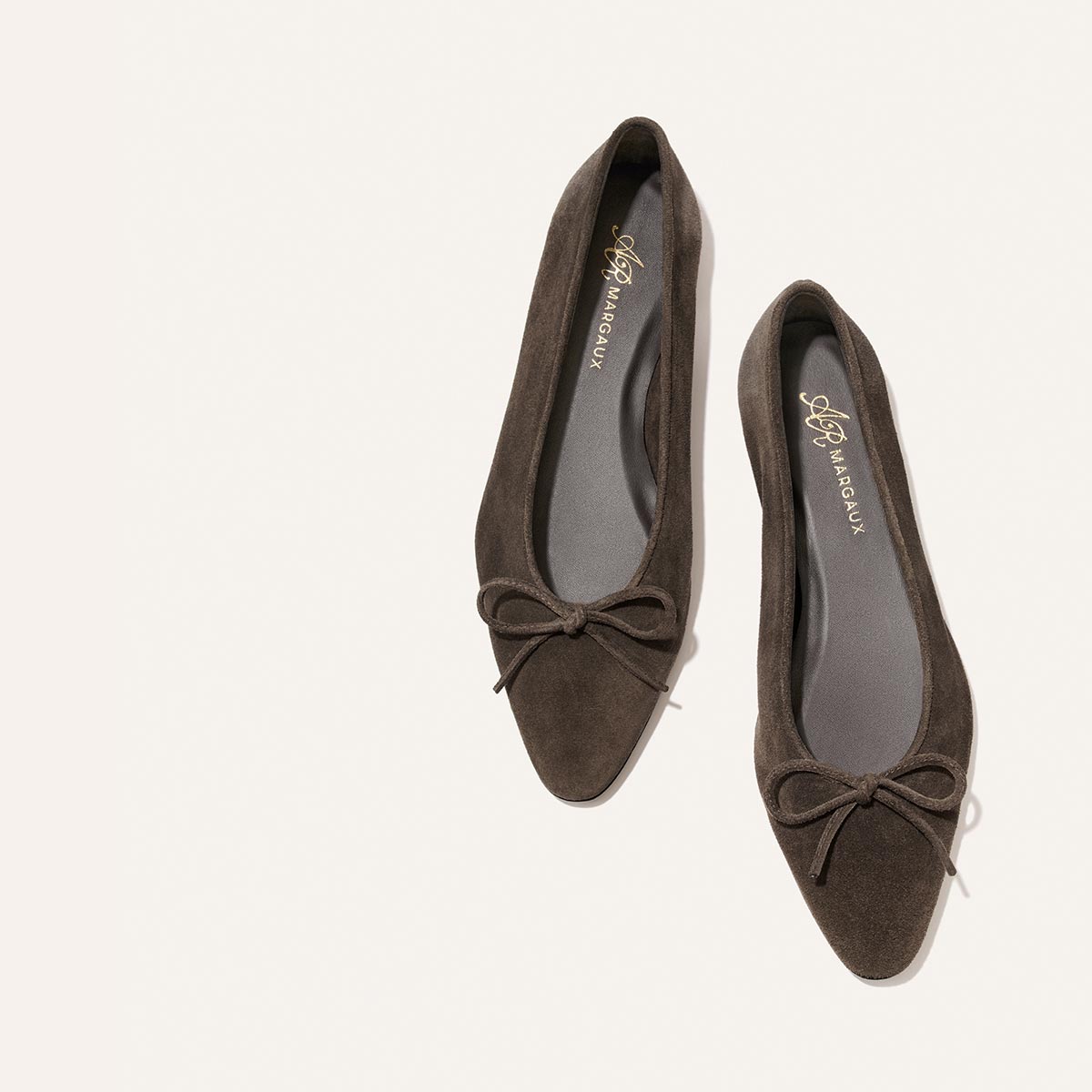 A pair of Margaux Alison Roman x Margaux: The Salamanca - English Brown Suede ballet flats with toe bows, arranged diagonally on a light background.