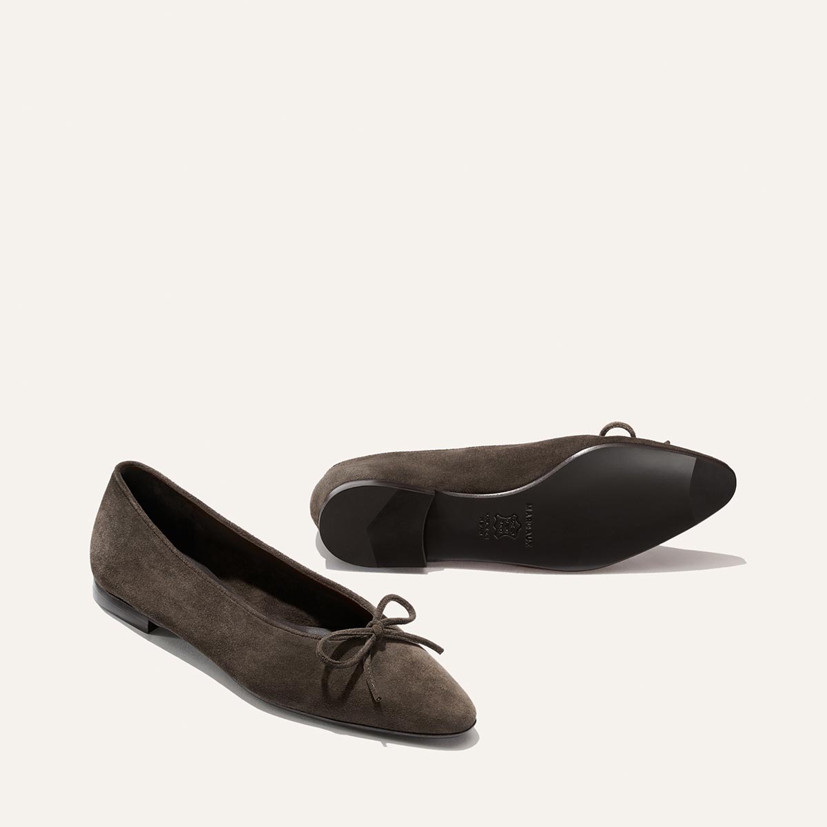 A pair of Margaux Alison Roman x Margaux: The Salamanca pointed-toe ballet flats in English Brown suede, featuring elegant bow accents; one shoe is upright while the other lies on its side to display the sole, both set against a plain light background.