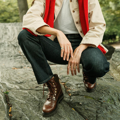 A person crouches on a rock outdoors, wearing dark jeans and the margaux-development Aspen Boot in Russet Calf. They pair it with a beige jacket, white shirt, and red scarf, surrounded by greenery; their head is out of frame.