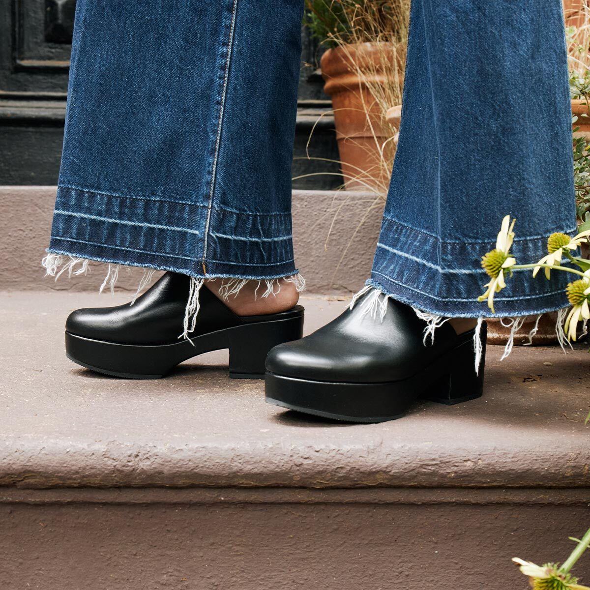 Wearing the Alison Roman x Margaux: The Atlantic Clog in Black Calf by Margaux with wide, frayed-hem jeans, standing on outdoor steps near potted plants and yellow flowers—a true Brooklyn street shoe vibe.