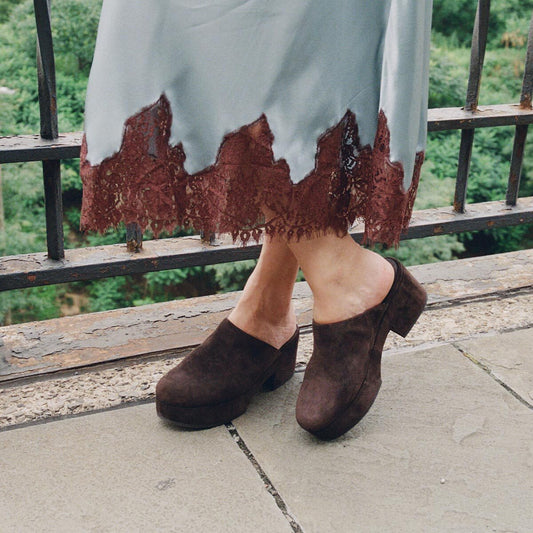 A person wearing a long skirt with lace trim and stylish Margaux Alison Roman x Margaux: The Atlantic Clog in Dark Chocolate Suede stands on a stone path next to a metal railing with greenery in the background.