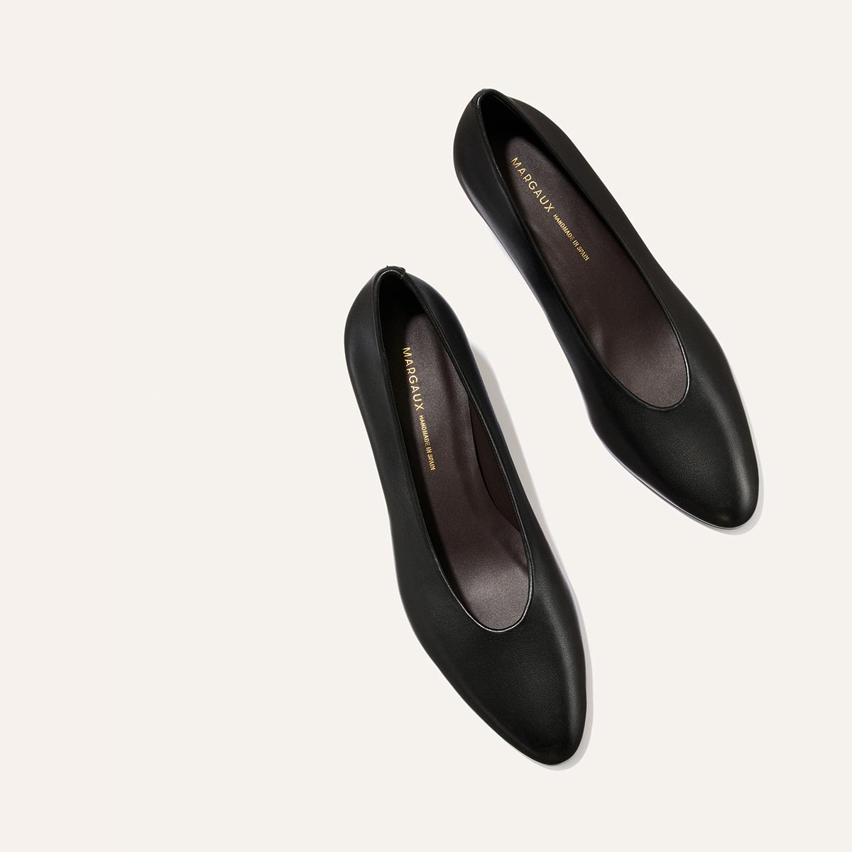 A pair of Margaux’s The Augusta Glove Pump in Black Nappa leather, featuring rounded toes and gold branding on the insoles, is displayed from above against a light beige background.