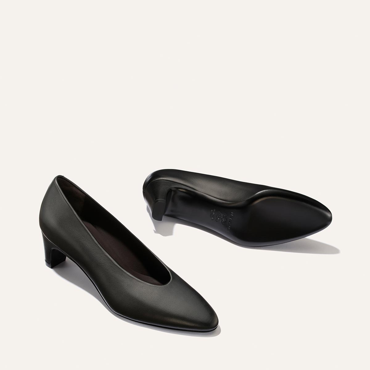 A pair of The Augusta Glove Pump - Black Nappa by Margaux is displayed on a simple light background. One shoe stands upright, while the other lies on its side to reveal the sole and low heel.