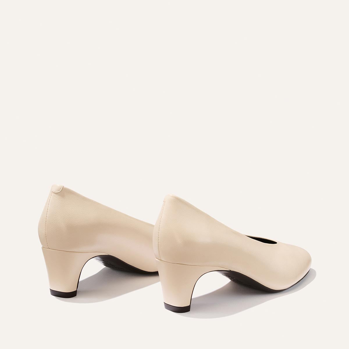 A pair of Margaux’s The Augusta Glove Pump in Bone Nappa—chic beige low-heeled women’s pumps with rounded toes—displayed on a plain white background and shown from both the back and side angles.