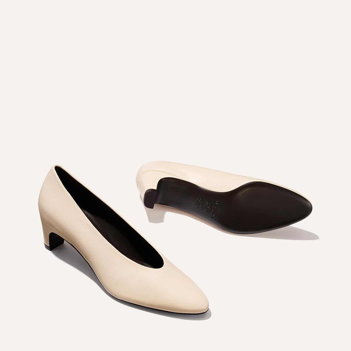 A pair of Margaux’s The Augusta Glove Pump in Bone Nappa—a beige low-heeled style with rounded toes—is displayed on a plain, light background. One shoe stands upright, while the other rests on its side, revealing the black sole and small block heel.